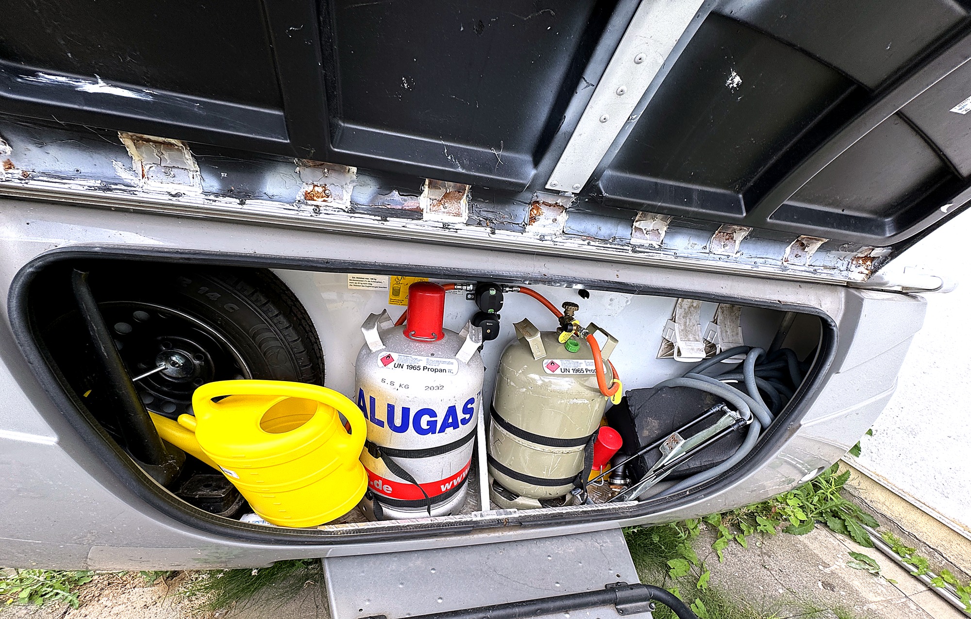 Gaskasten mit zwei Gasflaschen, Regler und Reserverad. Die Klappenkante zeigt Rostansatz – so wie es ist.
